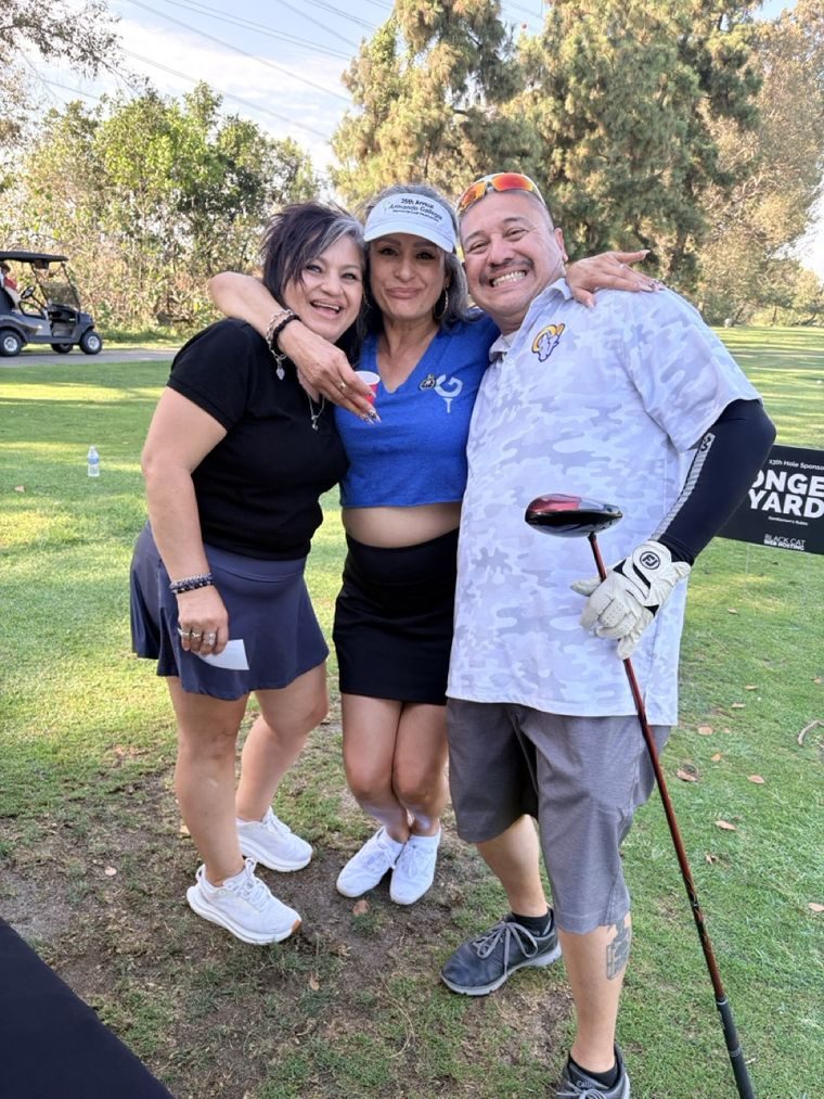 Three guests at the 13th hole, one wearing a 25th Annual Armando Gallegos visor, with the Longest Yard sign behind them.